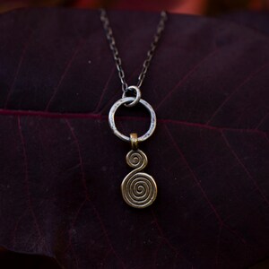 Puede incluir: Un collar de cadena de plata con un colgante circular de plata y un colgante de espiral dorado. El collar est&aacute; sobre una hoja de color rojo oscuro.