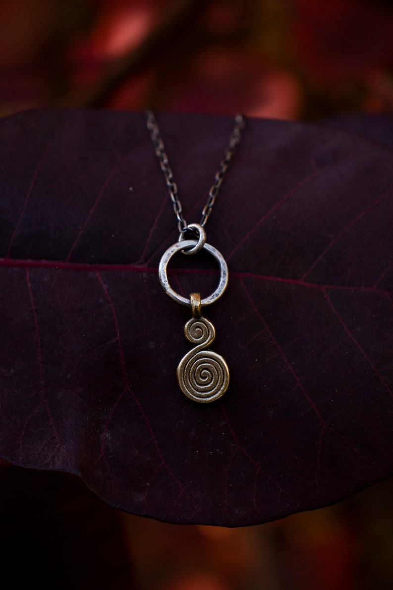 Puede incluir: Un collar de cadena de plata con un colgante circular de plata y un colgante de espiral dorado. El collar est&aacute; sobre una hoja de color rojo oscuro.
