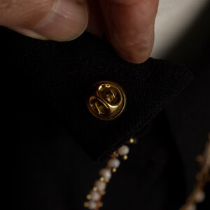 May include: A gold-coloured pin with a round head, attached to a black fabric garment.