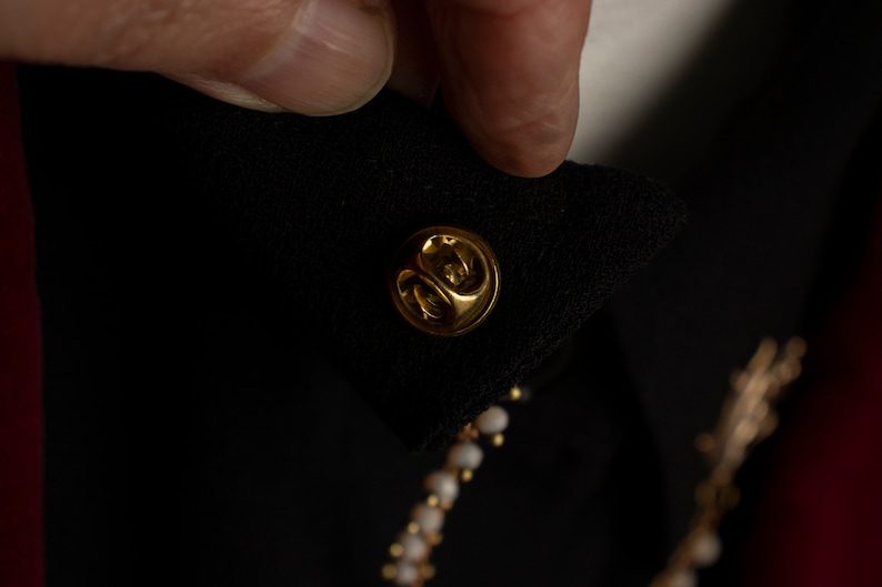 May include: A gold-coloured pin with a round head, attached to a black fabric garment.