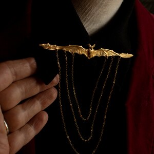 Pu&ograve; includere: Un fermaglio a forma di pipistrello dorato con un dettaglio a catena. Il fermaglio &egrave; indossato su una camicia nera con una giacca rossa.
