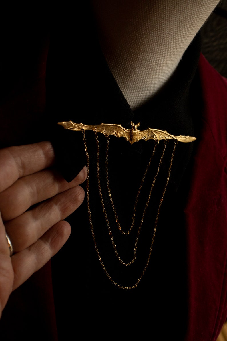 Pu&ograve; includere: Un fermaglio a forma di pipistrello dorato con un dettaglio a catena. Il fermaglio &egrave; indossato su una camicia nera con una giacca rossa.