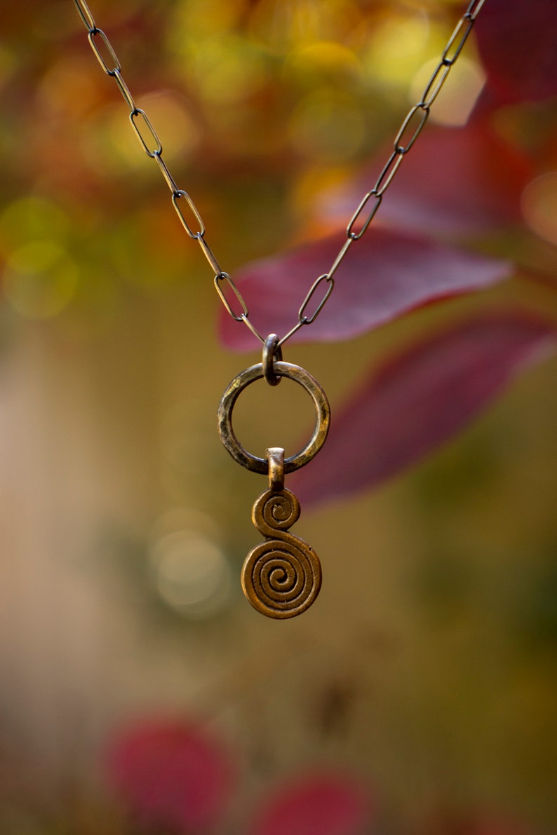 Puede incluir: Un collar de oro con un colgante en espiral y un dije circular. El collar est&aacute; colgado sobre un fondo borroso de follaje oto&ntilde;al.
