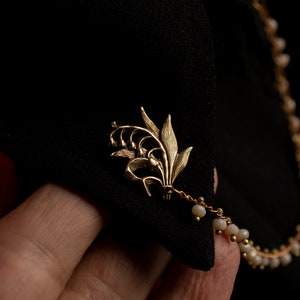May include: A gold lily flower brooch with a delicate chain and white beads attached.