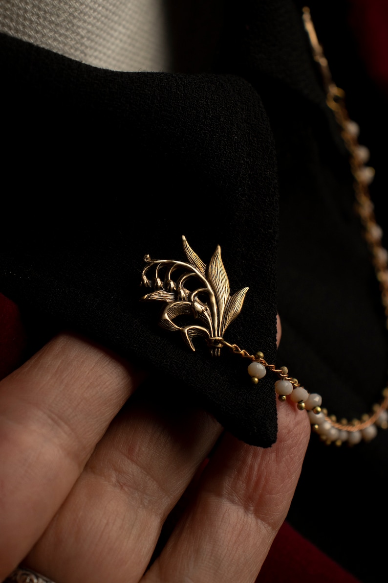 May include: A gold lily flower brooch with a delicate chain and white beads attached.