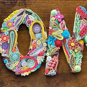 Può includere: Un'insegna "HOME" colorata e artigianale realizzata con la tecnica del quilling di carta. Ogni lettera è decorata con vivaci spirali di carta, fiori e altri abbellimenti nei toni del rosso, giallo, blu e verde. L'insegna è posta su uno sfondo di legno scuro.