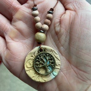 May include: A beige ceramic pendant with a tree of life design hangs from a brown leather cord with wooden beads. The pendant has a light blue and white swirl pattern.