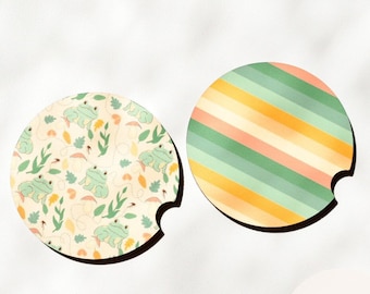 May include: Two round, cork-backed coasters with a cut-out for a beverage. One coaster features a green, yellow, and orange striped pattern. The other coaster features a pattern of green frogs, leaves, and hearts on a cream background. The coasters are labeled "Lexus Nalene Unique Gifting".