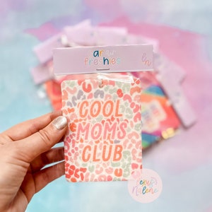 May include: A pink and white car air freshener with a leopard print design and the text "Cool Moms Club".