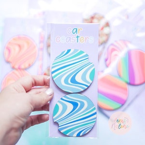 Pode incluir: Dois porta-copos para carro com um padr&atilde;o de redemoinho azul e branco. Os porta-copos est&atilde;o em uma manga de pl&aacute;stico transparente com o texto "car coasters" na embalagem.