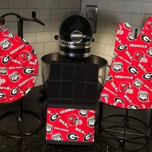 May include: A set of red and white kitchen accessories featuring the University of Georgia Bulldogs logo. The set includes a pot holder, a hot pad, and an oven mitt. The accessories are all made of fabric and have a quilted design.