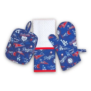 May include: A blue and red kitchen towel with a white background and a red border. The towel has a baseball theme with the words "Dodgers" and "Los Angeles" printed on it. The towel is accompanied by a blue and red oven mitt and a blue and red pot holder, both with the same baseball theme.