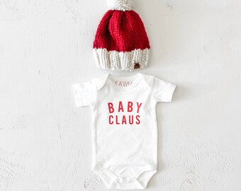 May include: A knitted baby Santa hat in red and white with a white bobble sits above a white baby bodysuit. The bodysuit has the words "BABY CLAUS" printed in red. The word "FAWN" is printed above the bodysuit.