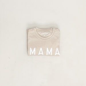 K&ouml;nnte beinhalten: Ein beiges T-Shirt mit dem Wort "MAMA" in wei&szlig; auf der Vorderseite.