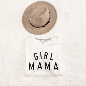 Peut inclure: Un t-shirt blanc avec le texte "GIRL MAMA" en lettres noires, associ&eacute; &agrave; un chapeau fedora beige avec un ruban noir. Le chapeau est plac&eacute; au-dessus du t-shirt sur une surface blanche textur&eacute;e. Le t-shirt porte la marque "FAWN md" sur le col.