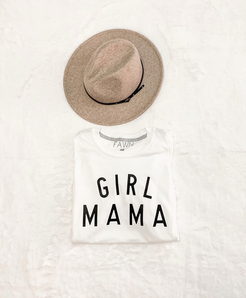 Peut inclure: Un t-shirt blanc avec le texte "GIRL MAMA" en lettres noires, associ&eacute; &agrave; un chapeau fedora beige avec un ruban noir. Le chapeau est plac&eacute; au-dessus du t-shirt sur une surface blanche textur&eacute;e. Le t-shirt porte la marque "FAWN md" sur le col.