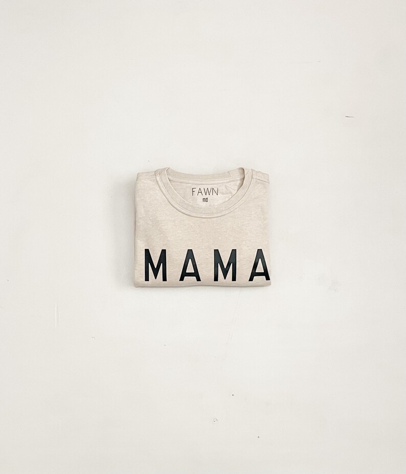 May include: Folded beige t-shirt with the word "MAMA" printed in bold black letters. The brand name "FAWN" is printed above the collar. A simple, neutral-toned garment.