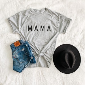 Peut inclure: Un t-shirt gris avec le mot "MAMA" en lettres noires, associ&eacute; &agrave; un short en jean et un chapeau fedora noir. Le t-shirt est nou&eacute; &agrave; la taille. Le short est en jean bleu. Le chapeau est rond avec un petit bord.