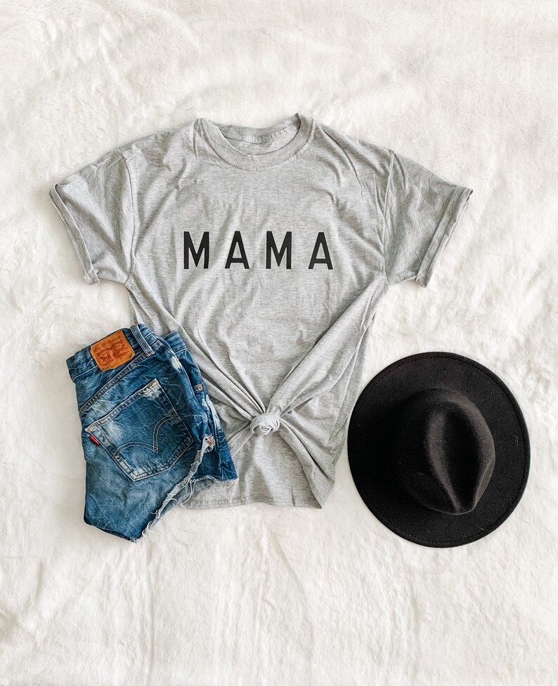 Peut inclure: Un t-shirt gris avec le mot "MAMA" en lettres noires, associ&eacute; &agrave; un short en jean et un chapeau fedora noir. Le t-shirt est nou&eacute; &agrave; la taille. Le short est en jean bleu. Le chapeau est rond avec un petit bord.