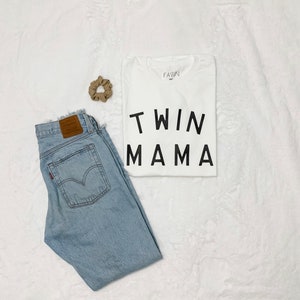 Peut inclure: Composition &agrave; plat avec un t-shirt blanc imprim&eacute; "TWIN MAMA" en noir, un jean bleu clair et un chouchou beige. Le jean a une &eacute;tiquette en cuir marron et une &eacute;tiquette rouge. Les articles sont dispos&eacute;s sur une surface blanche textur&eacute;e.