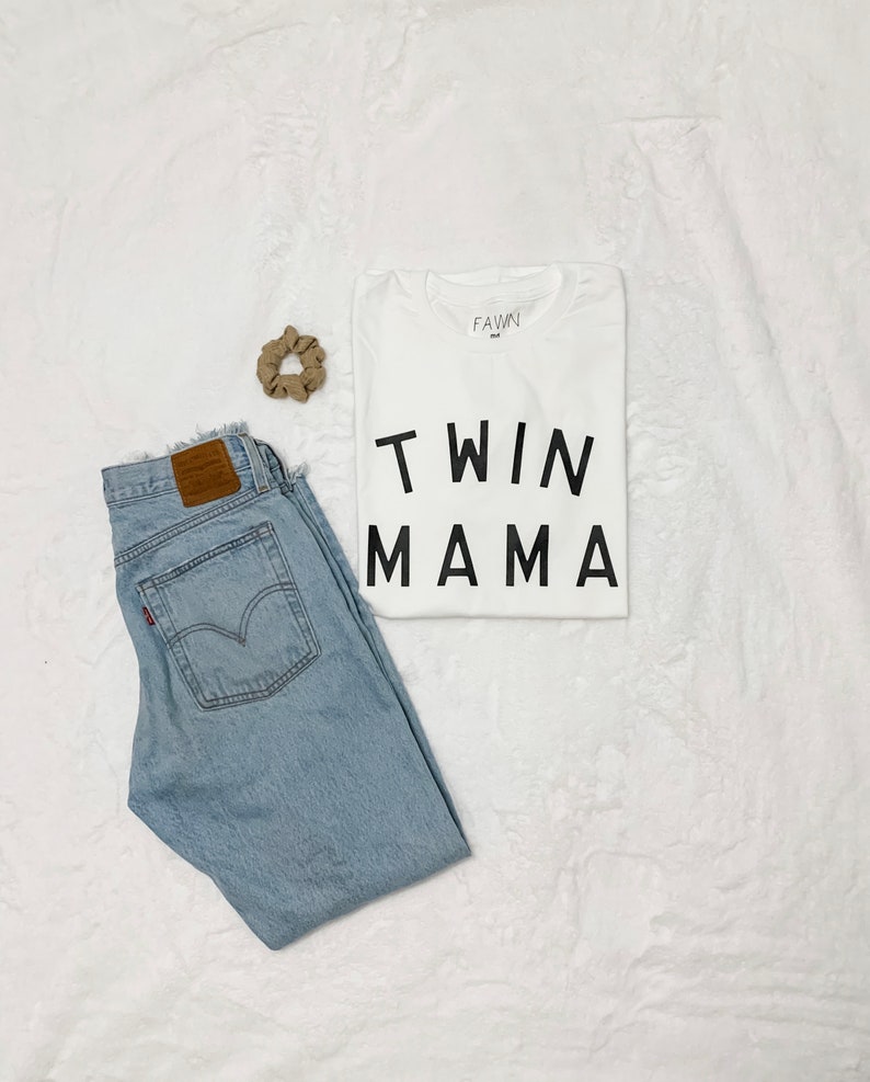 Peut inclure: Composition &agrave; plat avec un t-shirt blanc imprim&eacute; "TWIN MAMA" en noir, un jean bleu clair et un chouchou beige. Le jean a une &eacute;tiquette en cuir marron et une &eacute;tiquette rouge. Les articles sont dispos&eacute;s sur une surface blanche textur&eacute;e.