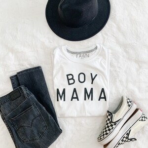 Peut inclure: Une tenue d&eacute;contract&eacute;e compos&eacute;e d'un chapeau fedora noir, d'un t-shirt blanc avec l'inscription "BOY MAMA", d'un jean d&eacute;lav&eacute; fonc&eacute; et de baskets &agrave; enfiler &agrave; carreaux. Le t-shirt porte la marque "FAWN" sur le col.