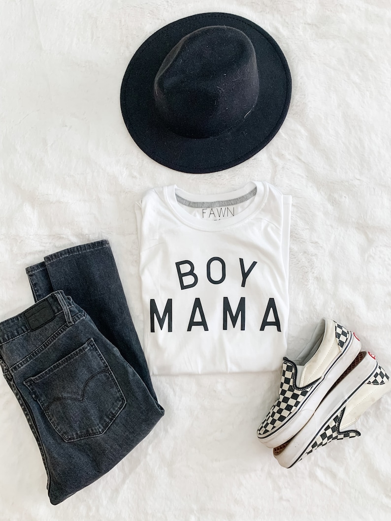 Peut inclure: Une tenue d&eacute;contract&eacute;e compos&eacute;e d'un chapeau fedora noir, d'un t-shirt blanc avec l'inscription "BOY MAMA", d'un jean d&eacute;lav&eacute; fonc&eacute; et de baskets &agrave; enfiler &agrave; carreaux. Le t-shirt porte la marque "FAWN" sur le col.