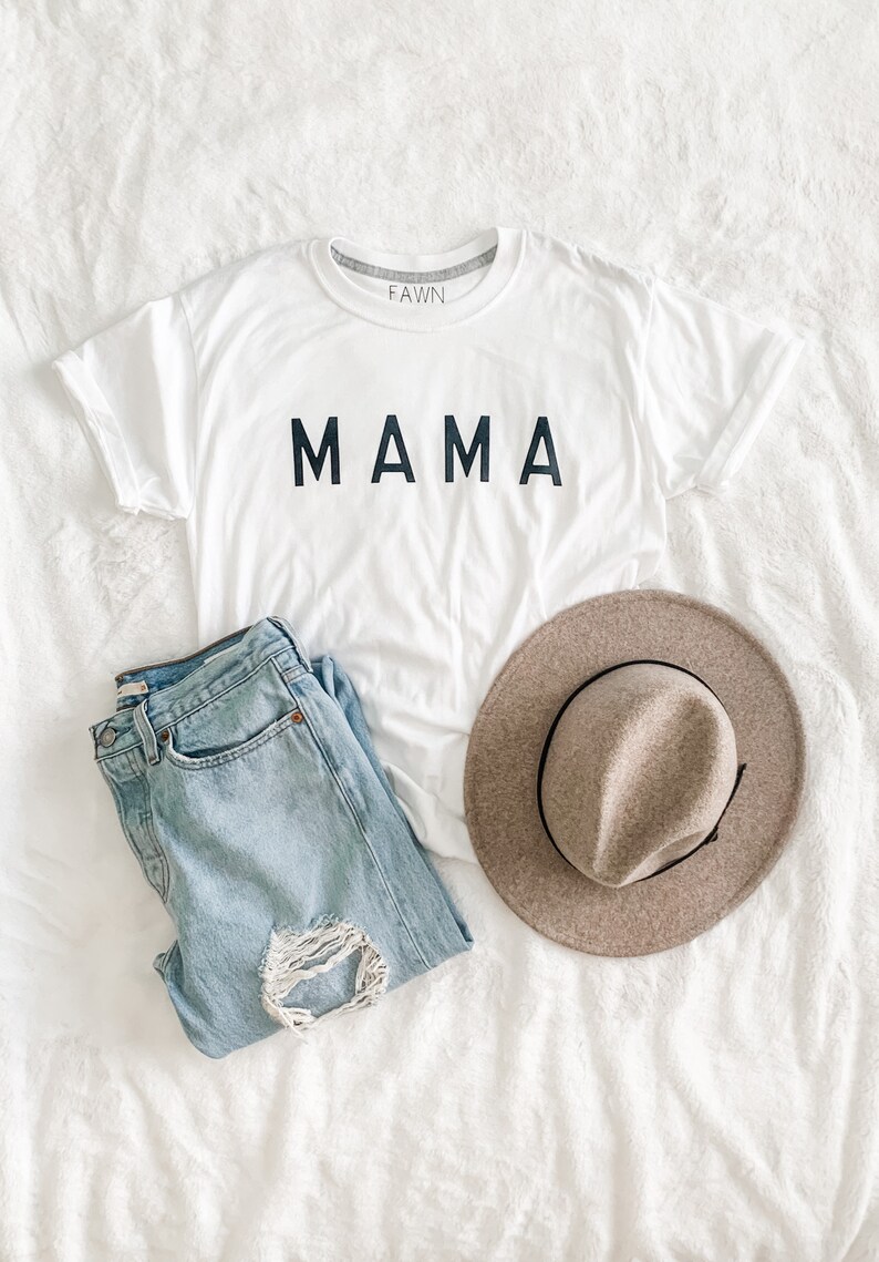 Peut inclure: Un t-shirt blanc avec le mot "MAMA" en lettres noires, un jean en denim bleu clair avec un genou d&eacute;chir&eacute; et un chapeau fedora beige avec un ruban noir, le tout dispos&eacute; sur une surface blanche textur&eacute;e.