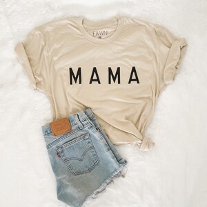 Peut inclure: Un t-shirt beige avec le mot "MAMA" imprim&eacute; en noir, associ&eacute; &agrave; un short en jean bleu clair. Le t-shirt est pli&eacute; et le short a un patch en cuir marron.