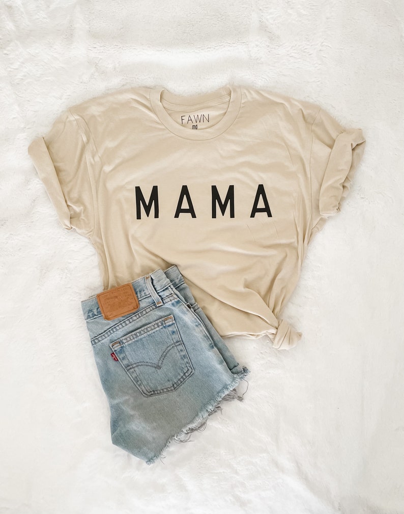 Peut inclure: Un t-shirt beige avec le mot "MAMA" imprim&eacute; en noir, associ&eacute; &agrave; un short en jean bleu clair. Le t-shirt est pli&eacute; et le short a un patch en cuir marron.