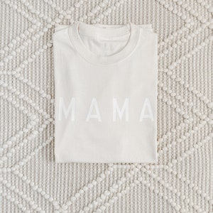 Peut inclure: Un t-shirt pli&eacute;, de couleur cr&egrave;me, avec le mot "MAMA" imprim&eacute; en grandes lettres majuscules blanches sur le devant. Le t-shirt est pos&eacute; sur un tapis textur&eacute; blanc cass&eacute; avec un motif en losange.