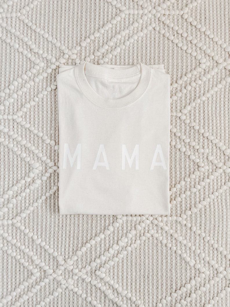 Peut inclure: Un t-shirt pli&eacute;, de couleur cr&egrave;me, avec le mot "MAMA" imprim&eacute; en grandes lettres majuscules blanches sur le devant. Le t-shirt est pos&eacute; sur un tapis textur&eacute; blanc cass&eacute; avec un motif en losange.