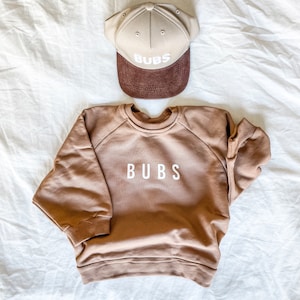 May include: A brown sweatshirt with the word "BUBS" embroidered on the front. A beige baseball cap with the same word embroidered on the front is on a white surface behind the sweatshirt.