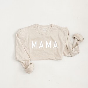 K&ouml;nnte beinhalten: Ein beiger Langarmsweater mit dem Wort "MAMA" in wei&szlig; auf der Vorderseite.