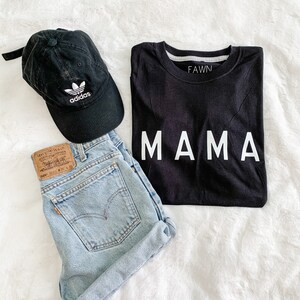 Peut inclure: Un ensemble d&eacute;contract&eacute; comprenant un t-shirt noir avec l'inscription "MAMA" en blanc, un short en jean d&eacute;lav&eacute; et une casquette de baseball Adidas noire. Le short a une &eacute;tiquette Levi's. La casquette arbore le logo Adidas.