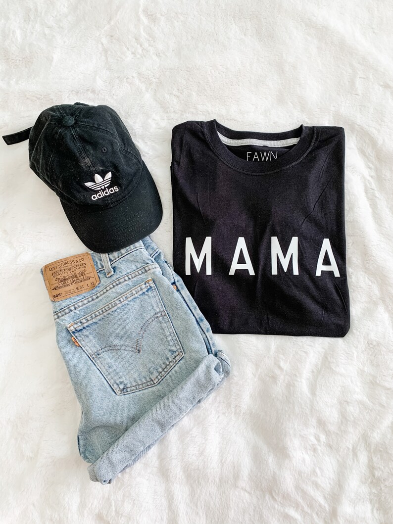 Peut inclure: Un ensemble d&eacute;contract&eacute; comprenant un t-shirt noir avec l'inscription "MAMA" en blanc, un short en jean d&eacute;lav&eacute; et une casquette de baseball Adidas noire. Le short a une &eacute;tiquette Levi's. La casquette arbore le logo Adidas.