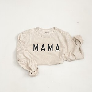 May include: A beige long-sleeved shirt with the word "MAMA" printed in bold black letters across the front. The shirt has a crew neck and the brand name "FAWN" is printed on the upper back. The sleeves are rolled up.