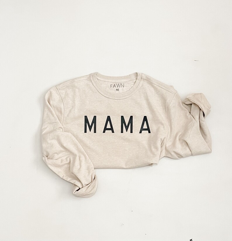 May include: A beige long-sleeved shirt with the word "MAMA" printed in bold black letters across the front. The shirt has a crew neck and the brand name "FAWN" is printed on the upper back. The sleeves are rolled up.