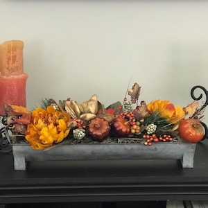 May include: A fall-themed centerpiece in a rectangular metal container. It features orange and yellow flowers, small pumpkins, berries, and greenery. Two pillar candles and a candle holder are on either side.