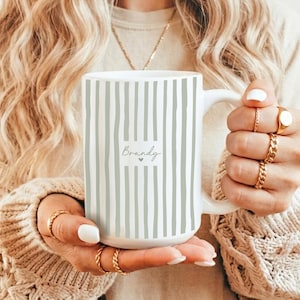 May include: White ceramic mug with vertical gray stripes. The mug features the name "Brandy" in a handwritten font with a small heart. The mug is held by a person wearing gold rings.