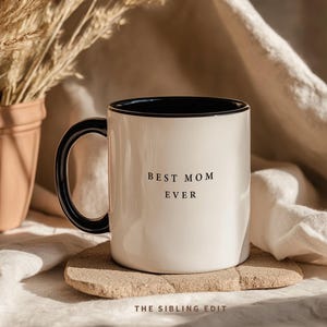 Op de afbeelding: Een witte keramische mok met een zwarte binnenkant en handvat. De mok heeft de tekst "BEST MOM EVER" in zwarte letters. Hij staat op een kleine, lichtgekleurde stenen onderzetter. De achtergrond omvat een terracotta pot met gedroogde planten en een beige doek.