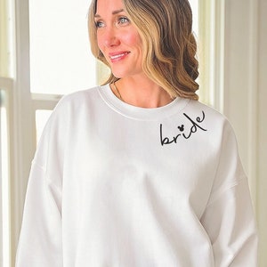 May include: White sweatshirt with the word "bride" in black script on the upper left chest. The word "bride" has two small hearts above the "i". The sweatshirt has long sleeves and a crew neck.
