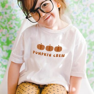 May include: A white short-sleeved t-shirt featuring three brown pumpkin illustrations and the text PUMPKIN CREW in brown lettering. The shirt is worn with mustard yellow leggings that have a small black polka dot pattern.