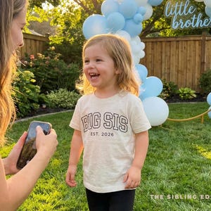 Puede incluir: Una ni&ntilde;a lleva una camiseta beige con el texto "BIG SIS EST. 2026" en negro. La ni&ntilde;a sonr&iacute;e en un jard&iacute;n, con globos azules y un letrero de "little brother" en el fondo.