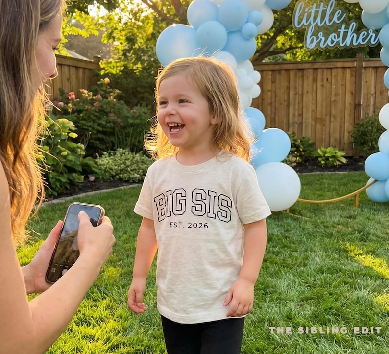 Puede incluir: Una ni&ntilde;a lleva una camiseta beige con el texto "BIG SIS EST. 2026" en negro. La ni&ntilde;a sonr&iacute;e en un jard&iacute;n, con globos azules y un letrero de "little brother" en el fondo.