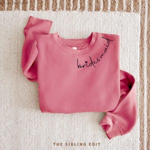 May include: A dusky pink sweatshirt with the word "bridesmaid" in black script across the chest. Two small hearts are placed before the word. The sweatshirt is folded on a white and beige striped surface.