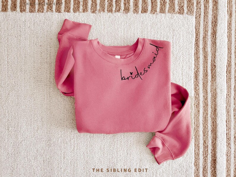 May include: A dusky pink sweatshirt with the word "bridesmaid" in black script across the chest. Two small hearts are placed before the word. The sweatshirt is folded on a white and beige striped surface.