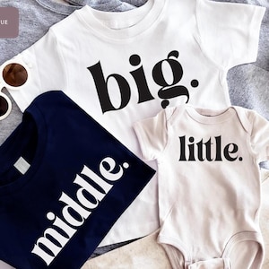 May include: A collection of children's apparel, including a white t-shirt with the word "big" in black, a navy blue t-shirt with "middle" in white, and a white onesie with "little" in black. Sunglasses are also visible.