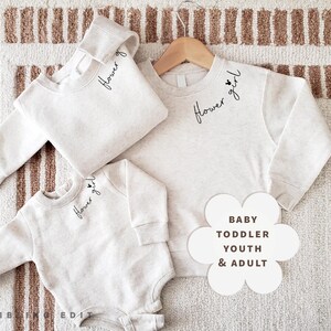 May include: Three light grey sweatshirts and a onesie with the words "Flower girl" in script. Sizes include baby, toddler, youth, and adult. The items are displayed on a striped rug.
