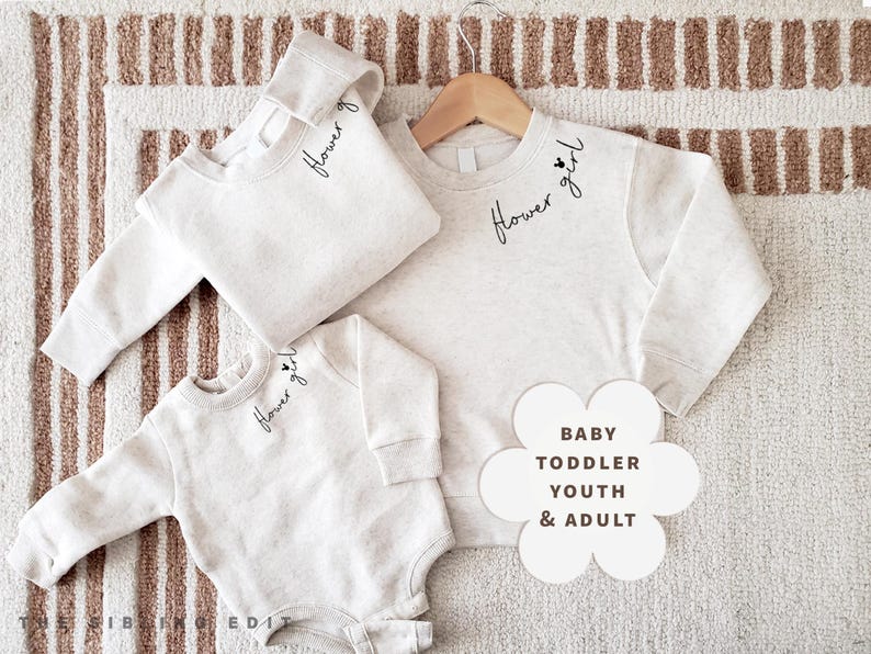 May include: Three light grey sweatshirts and a onesie with the words "Flower girl" in script. Sizes include baby, toddler, youth, and adult. The items are displayed on a striped rug.